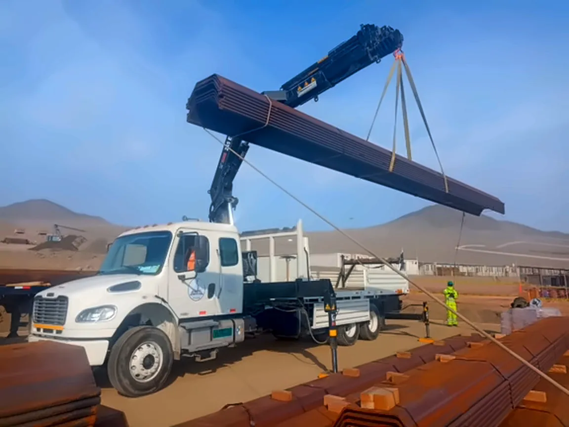 Camión grúa articulado izando un paquete de tuberías en obra